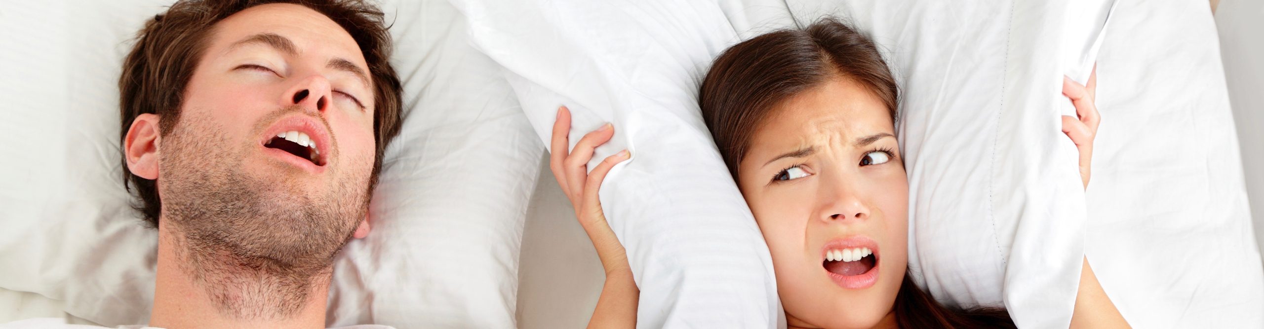 Woman covering her ears with pillows beside a sleeping man